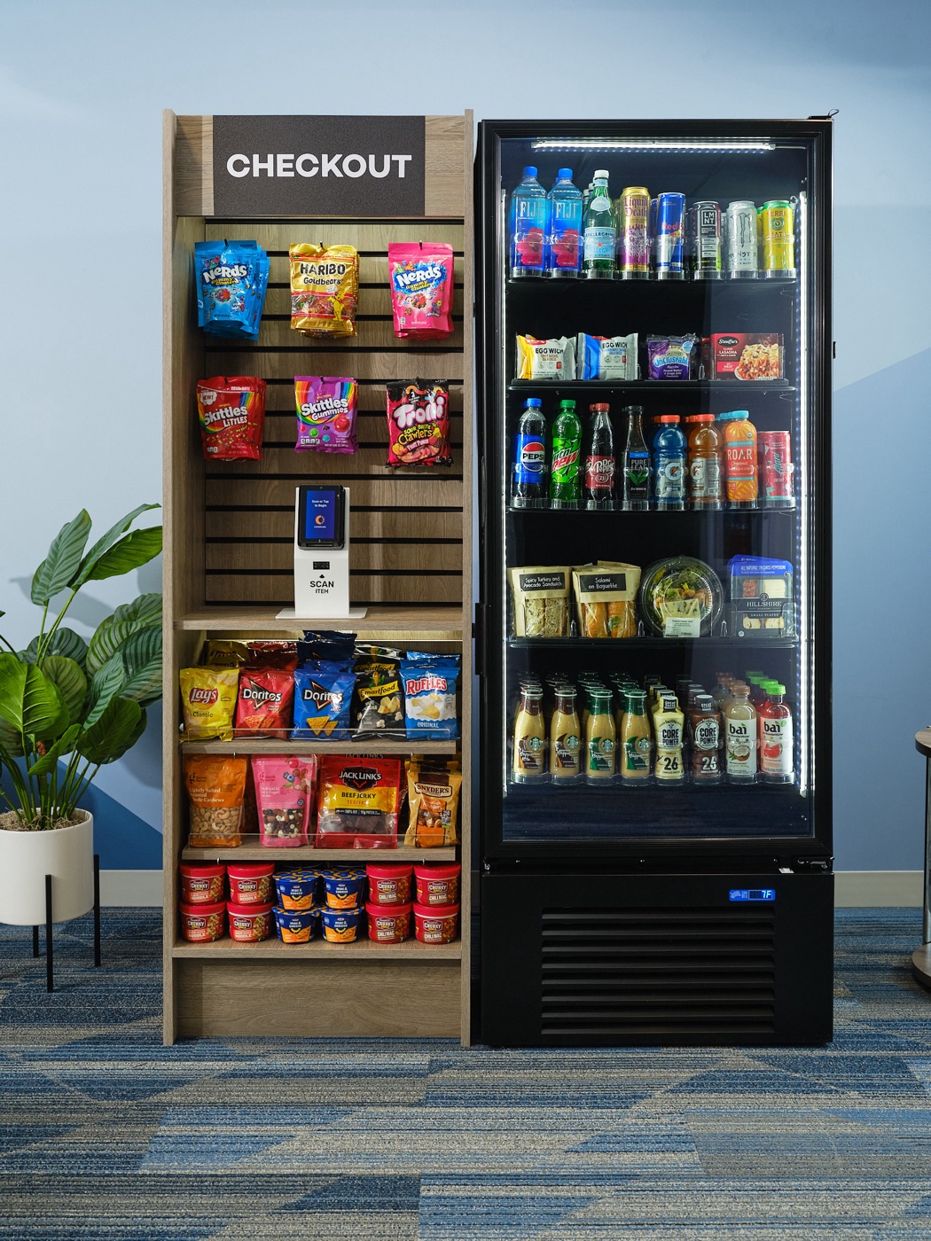 A micro market in a workplace break room: an open snack shelf with a self-checkout kiosk beside a glass-door cooler stocked with drinks, sandwiches and fresh meals