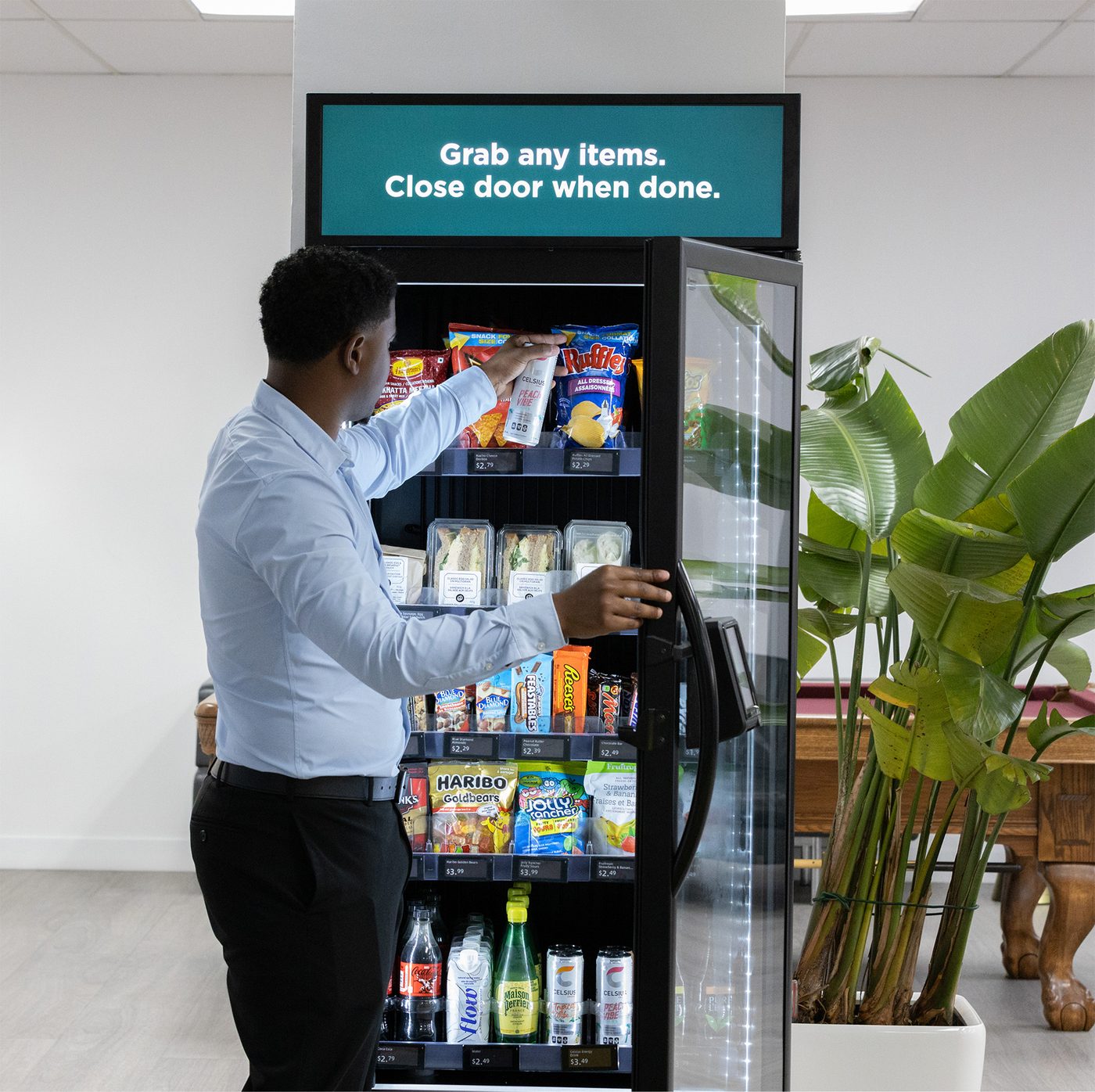 An employee reaching into a glass-door smart cooler stocked with snacks, sandwiches and cold drinks in a workplace break area