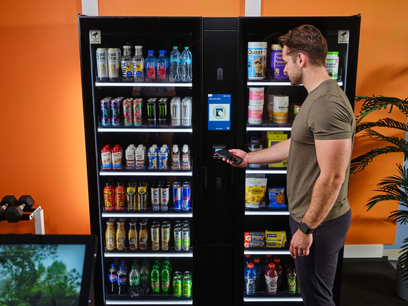 A gym member paying by phone at a smart vending unit stocked with protein shakes, energy drinks and recovery snacks, with dumbbells racked nearby