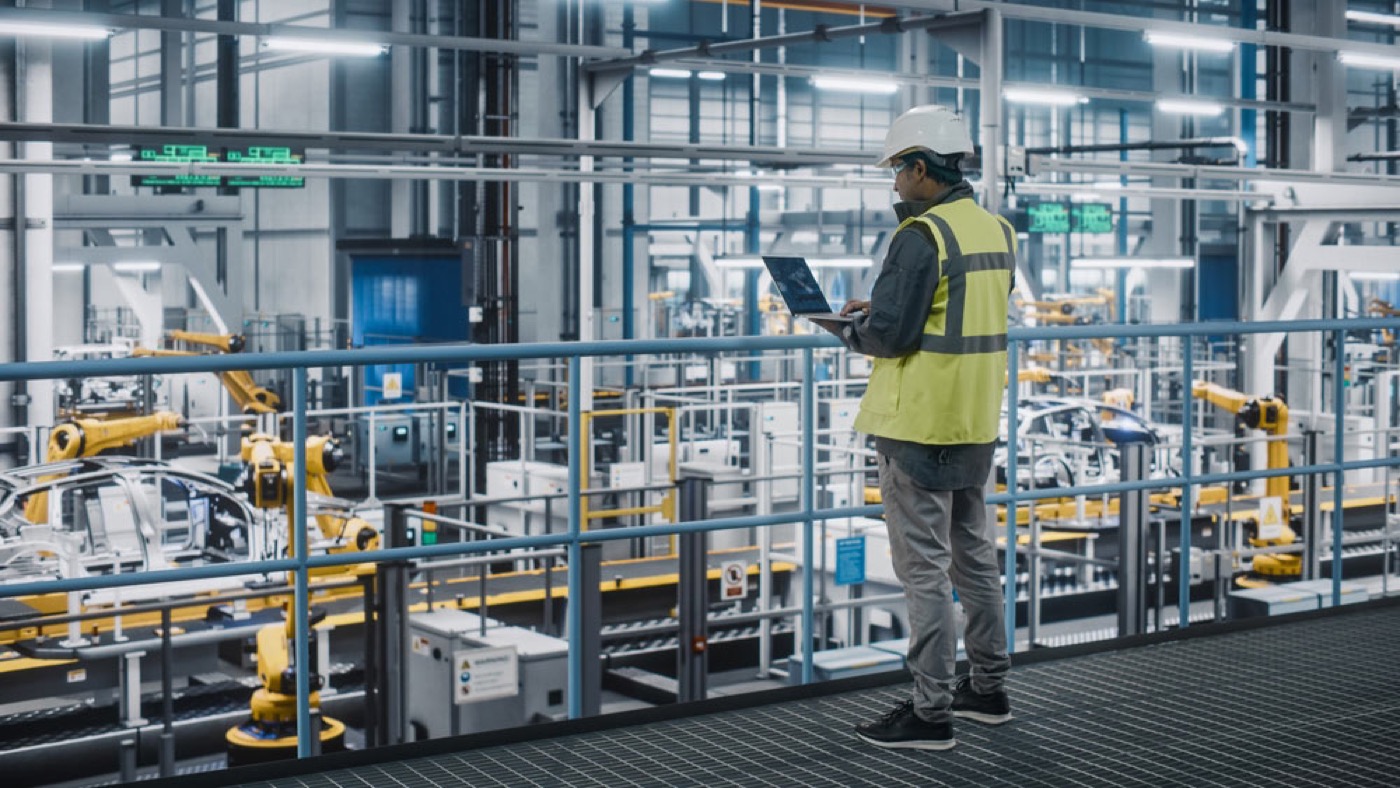 An engineer in a hard hat and high-visibility vest on a walkway above an automated production floor lined with robotic arms
