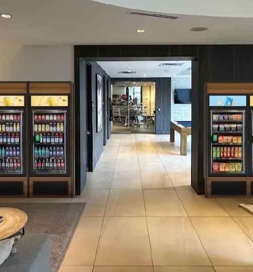 Coolers and snack displays flanking a corridor in an apartment community common area, available to residents around the clock